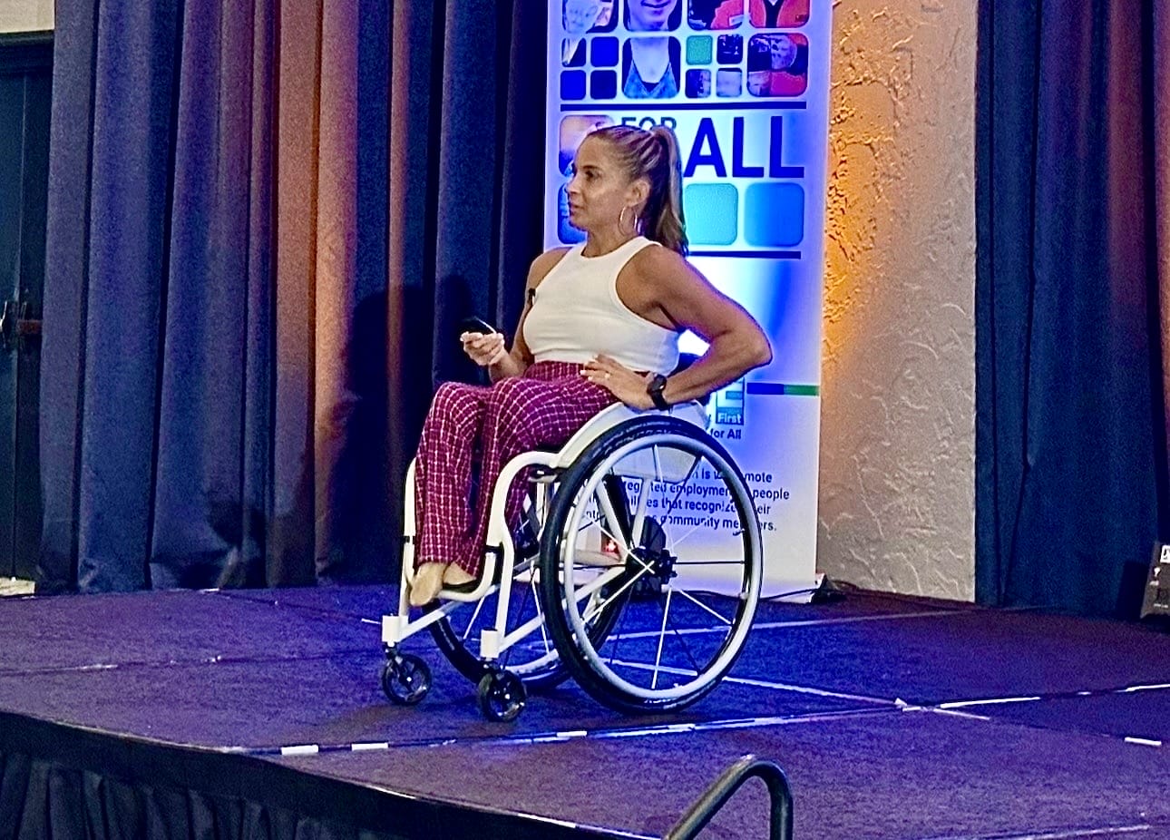 Alycia wearing red and white holding a remote control on stage speaking