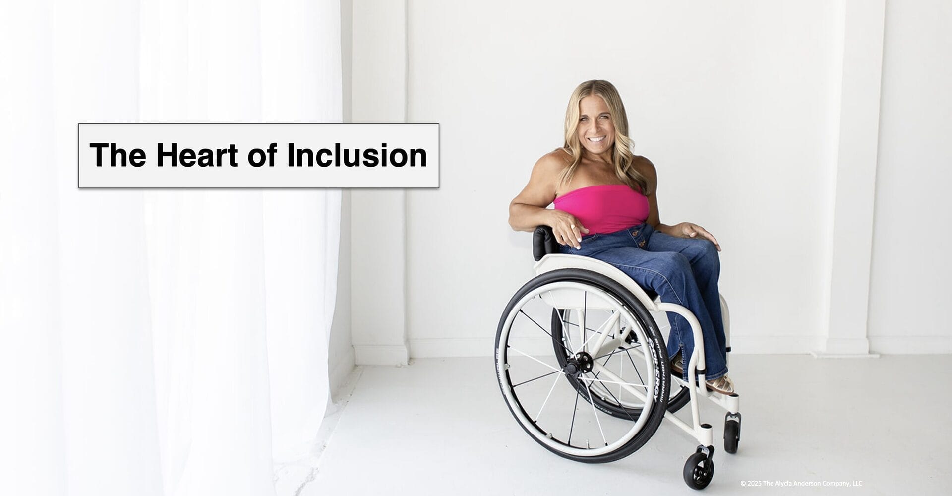 Alycia sitting in front of a white curtain in her wheelchair wearing pink and blue with "the heart of inclusion written next to her