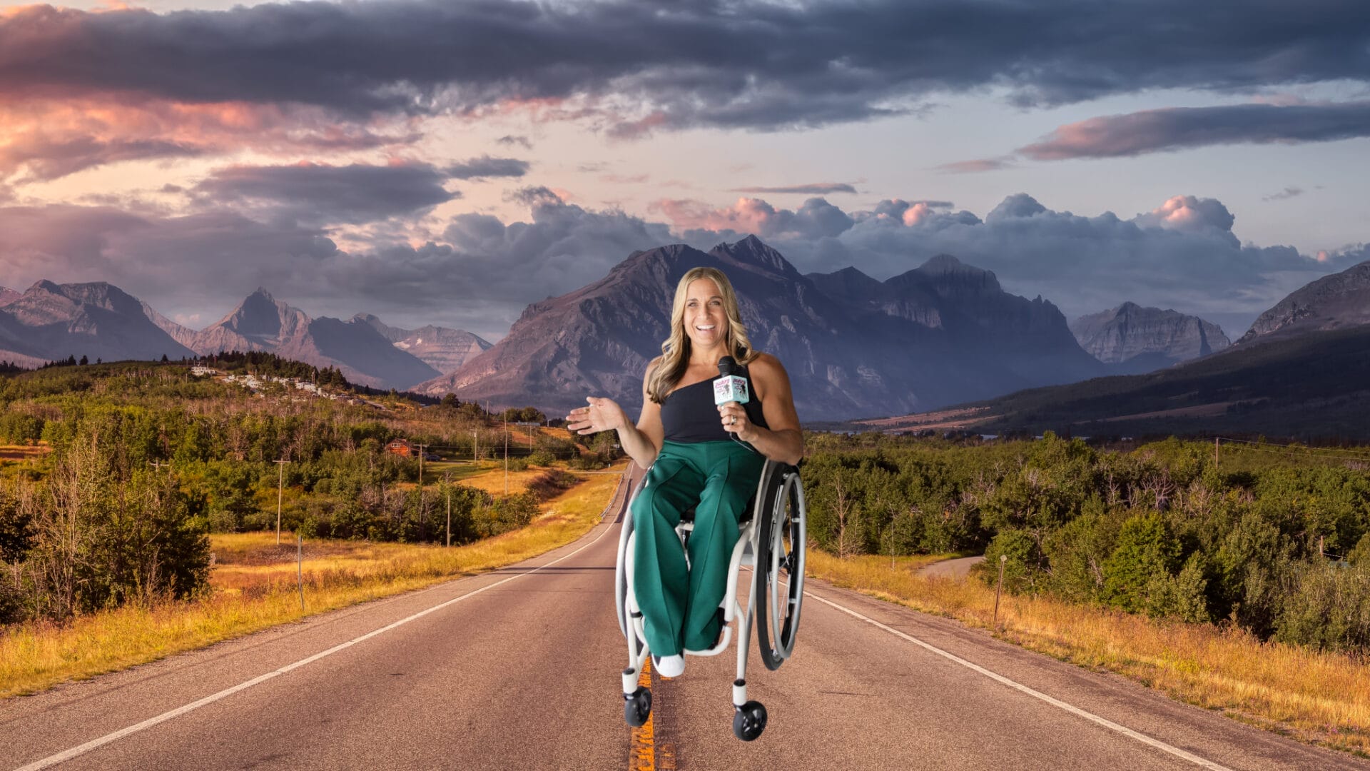 alycia sitting in the middle of an empty country road in her wheelchair holding her right hand out and holding a microphone with the other there mountains in the background