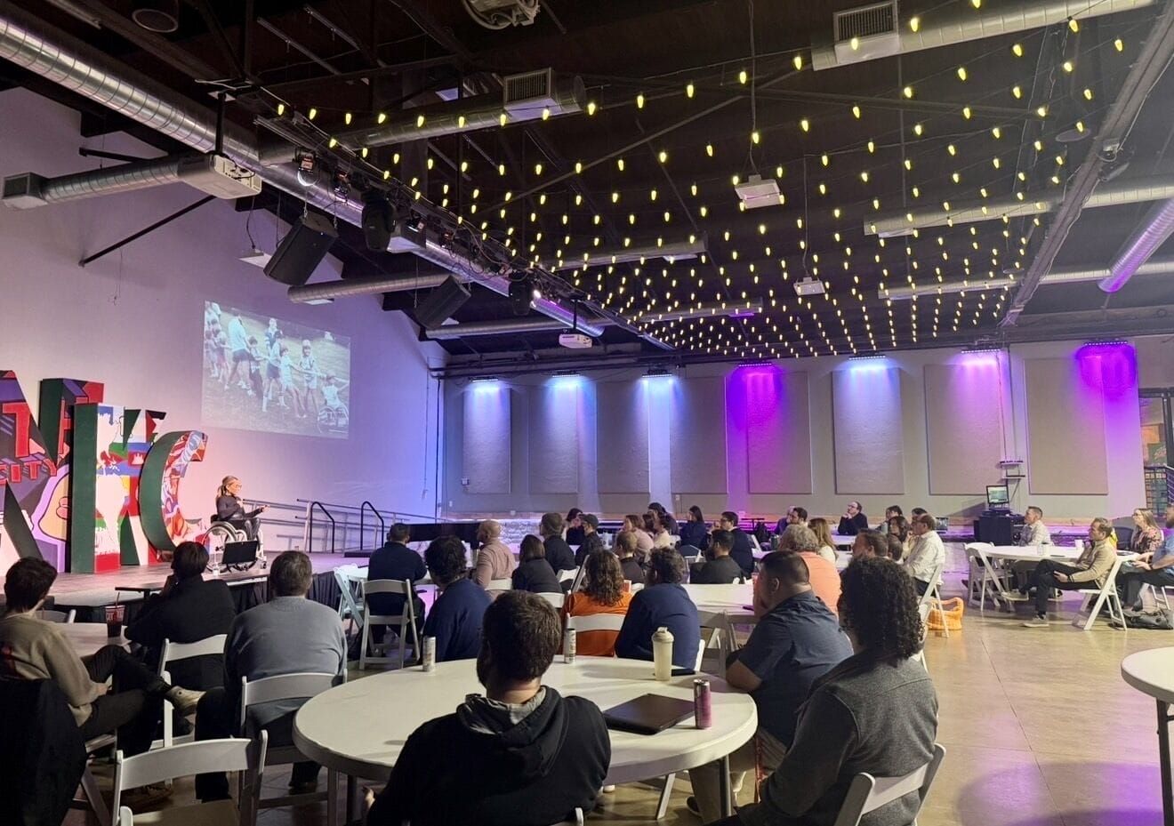 Wide view of Alycia Anderson speaking from her wheelchair on stage to an audience seated at round tables under string lights