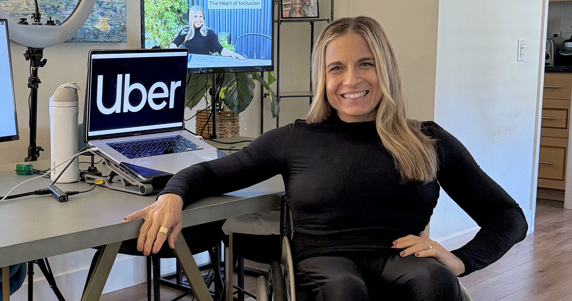 alycia sitting in her wheelchair resting one arm on her desk and one arm on her hip smiling with computer screens behind her one with the uber logo on the display