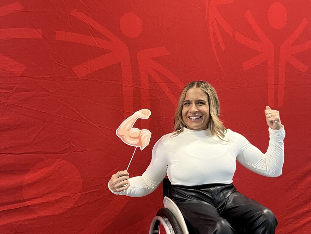 alycia flexing in front of a red special olympics marketing banner holding a arm flexing on a stick while sitting in her wheelchair and flexing with her other arm.