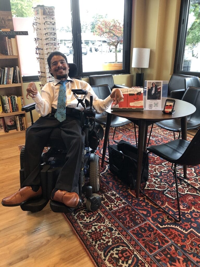 victor sitting in his power wheelchair at a book signing wearing a dress shirt tie slacks and dress shoes next to a table showcasing his book