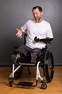 adam keys sitting in his wheelchair wearing a white t-shirt with his hand spread out wide smiling a big grin wearing a proshetic on his right arm and both legs