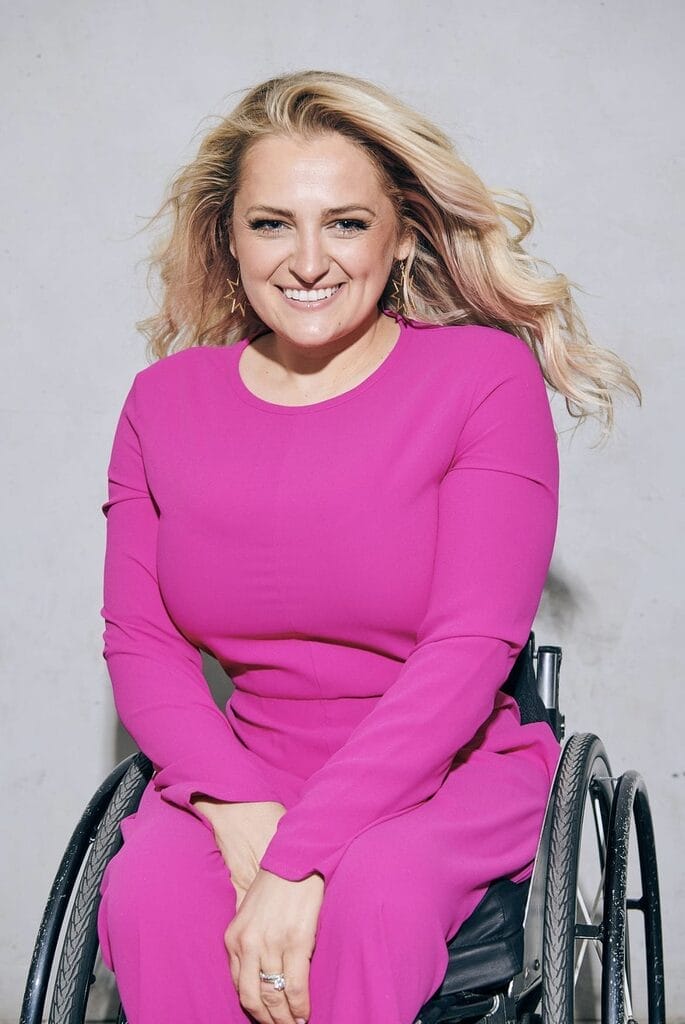 smiling blonde woman ali stroker in a bright pink outfit sits in her wheelchair against a light gray studio background